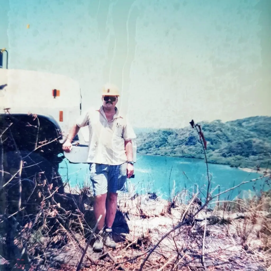 A young M J Harper standing in front of an industrial truck on a hillside overlooking the bay of Jonesville Point on Roatan in the Bay Islands