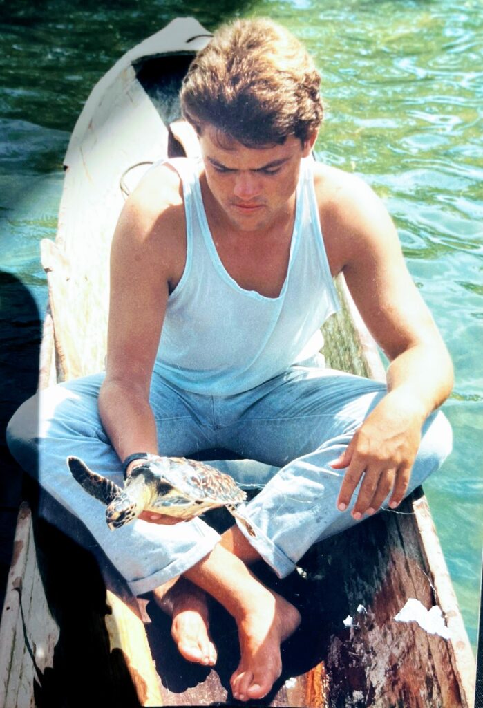 Photo of a young M J in the 1980's sitting in a canoe called the Dragon, holding a small turtle, Roatan history, Roatan stories